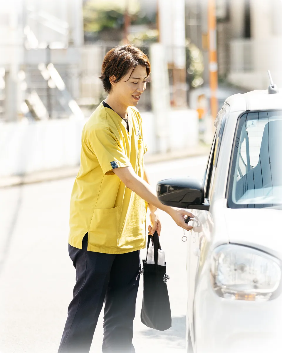 明るい黄色の制服を着た介護士が、晴れた日に白い車に乗り込もうとしている。