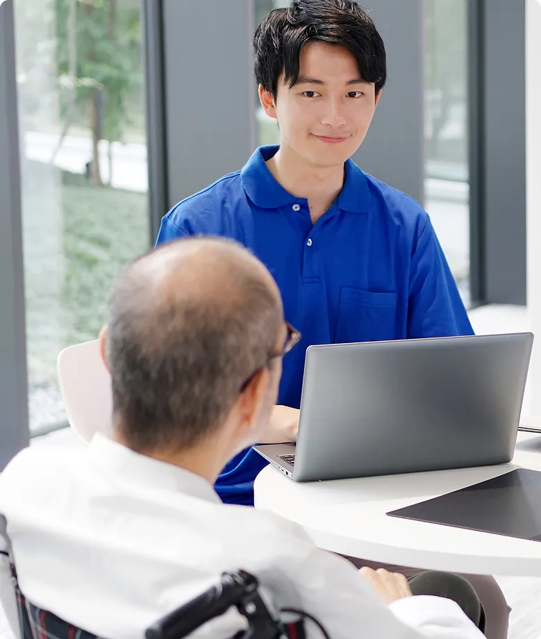 笑顔の青いポロシャツの男性が、白衣の年配の男性にデジタルサポートを提供している様子。