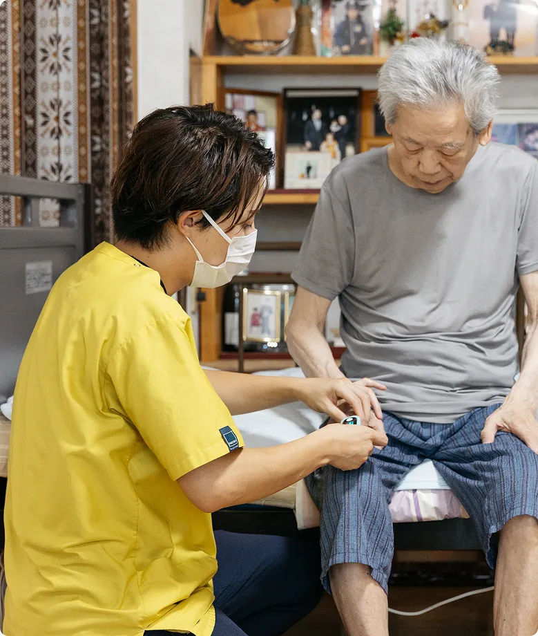介護士が明るい黄色のシャツを着て、高齢男性の指先で血中酸素飽和度を測定している様子。