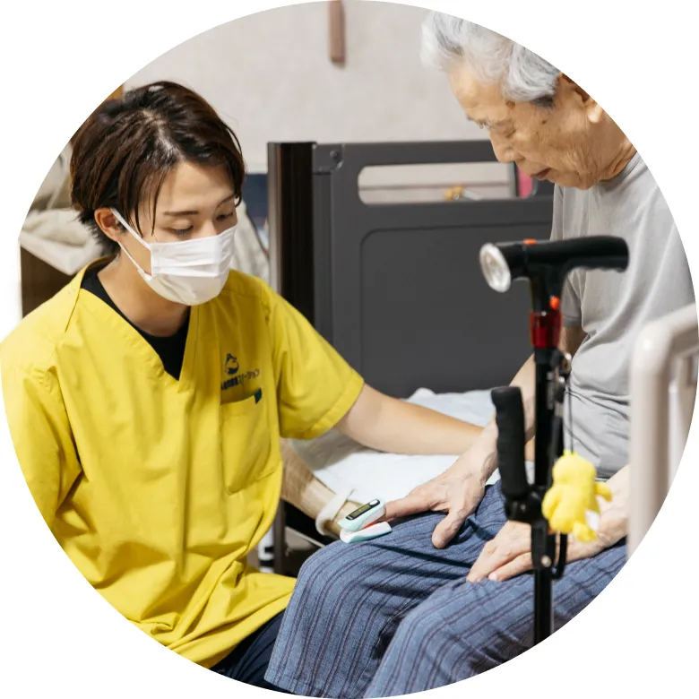 マスクをした若い介護士が、高齢男性の指にパルスオキシメーターを装着し、健康状態を確認している様子。