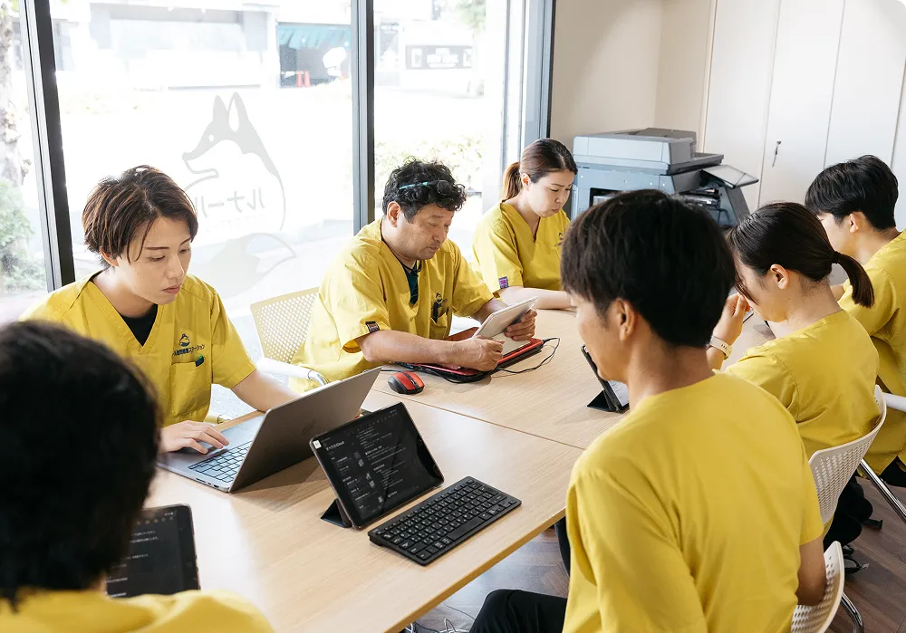 黄色いユニフォームを着たスタッフがタブレットやノートPCを使い会議を行っている様子