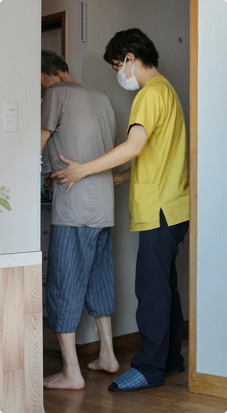 マスクをした介護者が、高齢者をそっと支えながら室内の出入りを手伝っている様子。