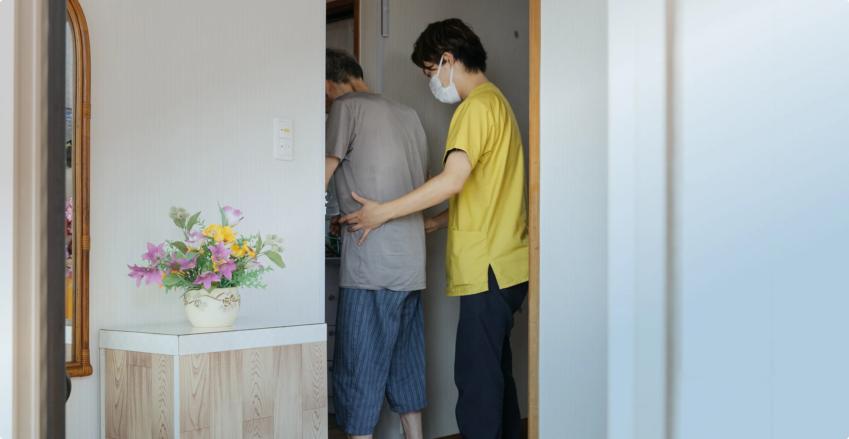 マスクをした介護者が、高齢者をそっと支えながら室内の出入りを手伝っている様子。